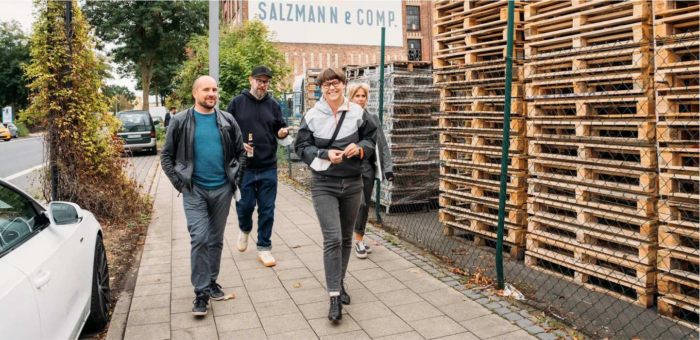 Vier Teammitglieder von machbar.de spazieren an einer Straße mit gestapelten Holzpaletten und einem Gebäude mit der Aufschrift "SALZMANN & COMP." vorbei.