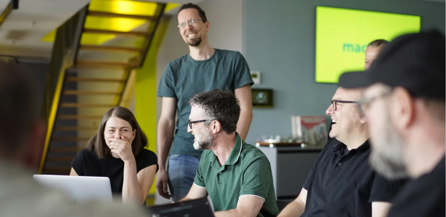 Ein Team von sechs Personen lacht und unterhält sich in einem Büro mit einer gelb-grünen Treppe und einem Bildschirm mit dem "machbar"-Logo.