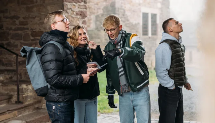 Vier junge Leute, drei Männer und eine Frau, stehen draußen und schauen sich um, während einer von ihnen auf etwas zeigt.